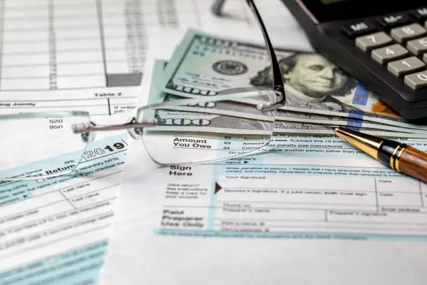 Photo of 1040 income tax return form 2019 with calculator, money, pen and focus through glasses on amount you owe. Concept of filing taxes, payment, refund, and April 15, 2020 tax deadline