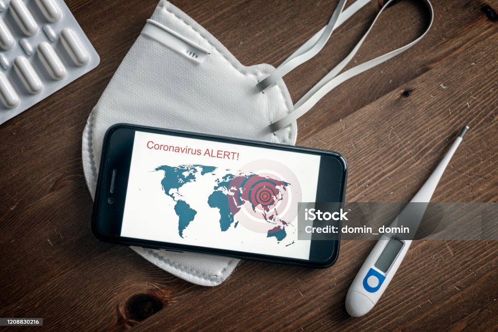 Coronavirus Covid-19 is spreading all over the world, mobile phone with world map is lying on the face mask Coronavirus (2019-nCoV) is spreading all over the world, mobile phone with world map and alert is lying on the white face mask amount pills and thermometre. Accidents and Disasters Stock Photo