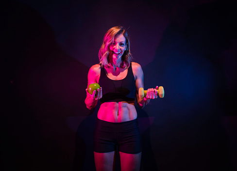 Front view of who is beautiful caucasian female exercising in front of colored background wearing sports bra who is smiling and being active with weights