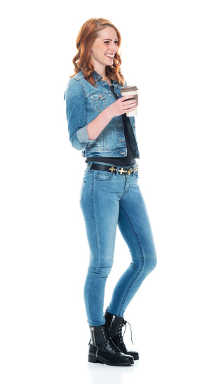 Profile view of aged 20-29 years old who is beautiful with redhead caucasian young women standing in front of white background wearing jeans who is cheerful and holding coffee cup