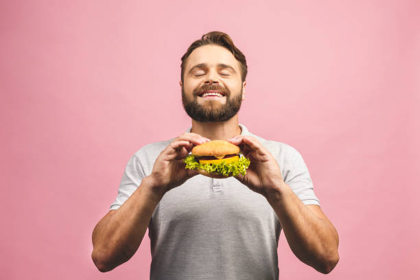 young man holding a piece of hamburger. bearded gyu eats fast food. burger is not helpful food. very hungry guy. diet concept. isolated over pink background. - burger hamburger large food imagens e fotografias de stock