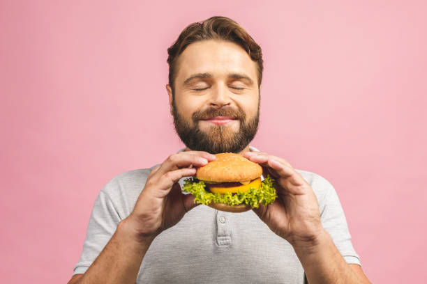 un giovanotto in possesso di un pezzo di hamburger. il gyu barbuto mangia fast food. l'hamburger non è cibo utile. un tipo molto affamato. concetto dietetico. isolato su sfondo rosa. - man eating foto e immagini stock