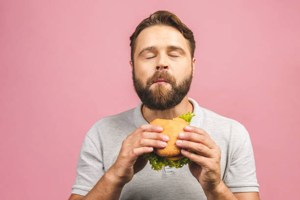Young man holding a piece of hamburger. Bearded gyu eats fast food. Burger is not helpful food. Very hungry guy. Diet concept. Isolated over pink background. Young man holding a piece of hamburger. Bearded gyu eats fast food. Burger is not helpful food. Very hungry guy. Diet concept. Isolated over pink background. greed photos stock pictures, royalty-free photos & images