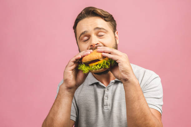 young man holding a piece of hamburger. bearded gyu eats fast food. burger is not helpful food. very hungry guy. diet concept. isolated over pink background. - burger hamburger large food imagens e fotografias de stock