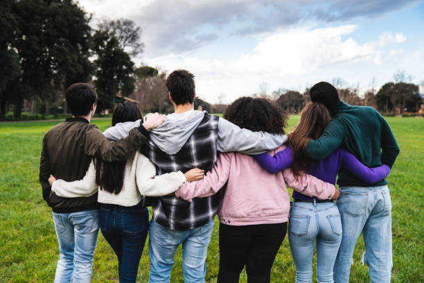 Group of six teenager friends embracing together at the park, rear view Group of six teenager friends embracing together at the park, rear view. Teamwork and cooperation concept with people together, sharing a common purpose. arm in arm stock pictures, royalty-free photos & images