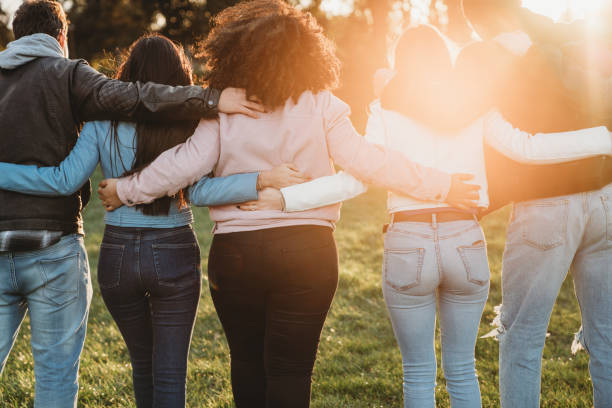 Group of six teenager friends embracing together at the park, rear view Group of six teenager friends embracing together at the park at sunset, rear view. Teamwork and cooperation concept with people together, sharing a common purpose. arm in arm stock pictures, royalty-free photos & images