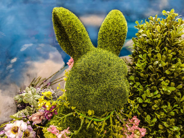 istock Decorative green topiary rabbit head with flowers around its neck and surrounded by greenery in front of blue sky-like background - Great for spring or Easter. 1208845274