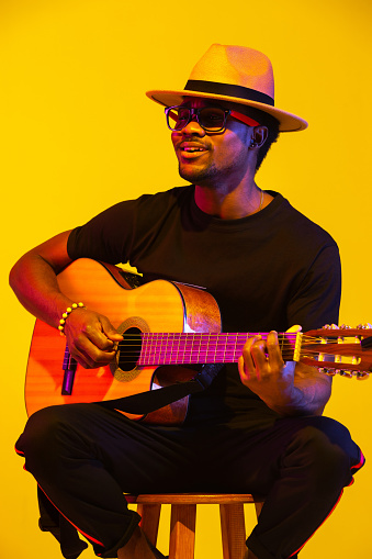 Young and joyful african-american musician playing guitar and singing on gradient orange-yellow studio background in neon light. Concept of music, hobby, festival. Colorful portrait of modern artist.