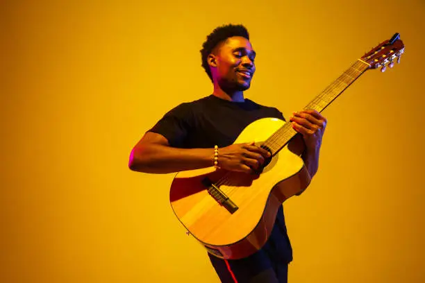 Photo of Young african-american musician singing, playing guitar in neon light