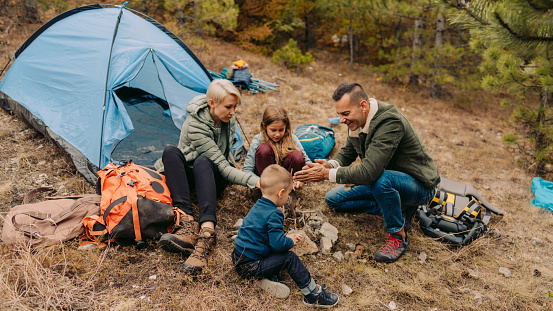 Photo of young family on a camping trip, gathered around the campfire in front of their tent // wide photo dimensions
