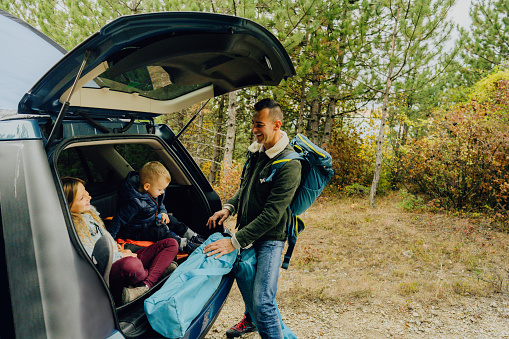 Photo of a single father with two kids, packing up a camping equipment in the trunk of a car