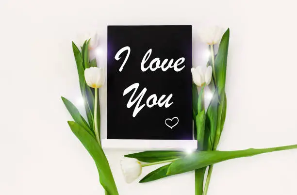 I love You card message sign on black chalkboard with tulip flowers on white background flat lay. Blackboard greeting text top view. Natural floral decoration with green leaves. Lovely romance banner