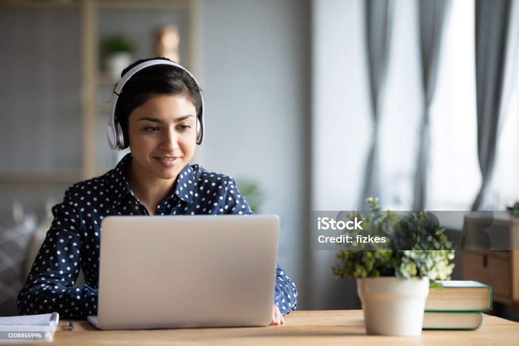 Millennial Indian girl in headphones using laptop at home Millennial Indian girl in wireless headphones sit at desk at home working on modern laptop, young ethnic woman in earphones browsing Internet shopping online or studying on computer in living room Internet Stock Photo