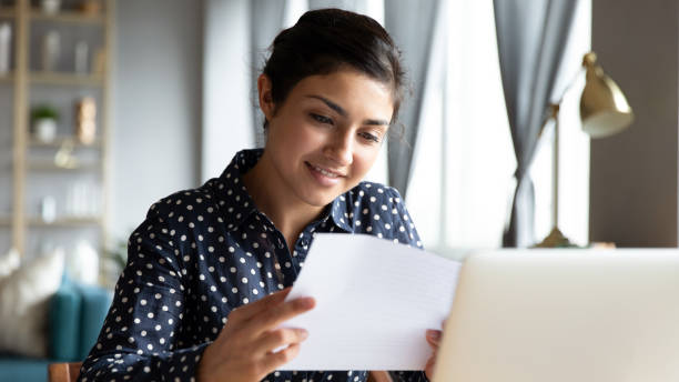 Happy Indian girl read good news in postal paper letter Happy millennial Indian girl sit at desk in living room reading good news in post paper correspondence, smiling young ethnic woman feel optimistic positive receive pleasant postal paperwork letter college acceptance letter stock pictures, royalty-free photos & images