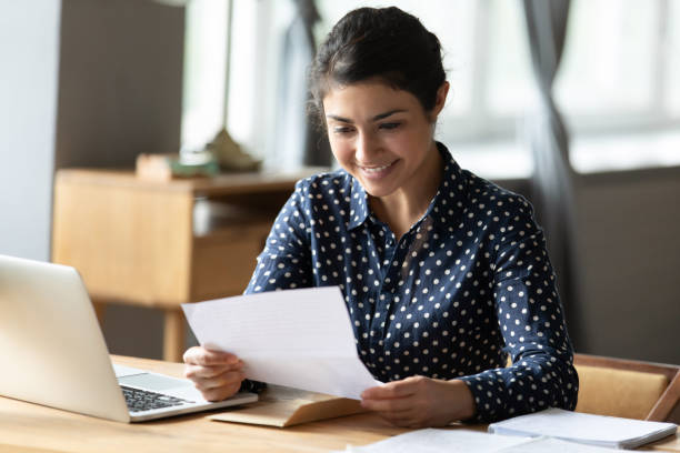 Smiling Indian woman read good news in paperwork correspondence Smiling Indian millennial girl sit at desk in living room read good news in paperwork correspondence, happy ethnic young woman distracted from computer work receive pleasant paper notice letter college acceptance letter stock pictures, royalty-free photos & images