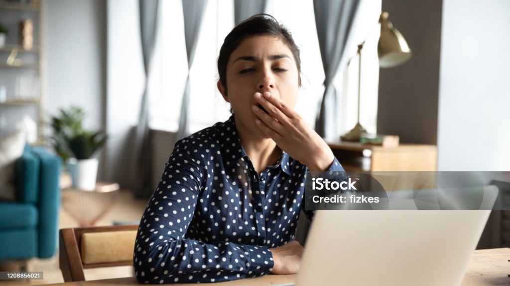 Tired ethnic woman yawn sigh at workplace Tired young Indian woman sit at desk in living room work at laptop yawn suffering from fatigue, exhausted millennial ethnic girl distracted from computer work job sigh feel asleep at workplace Working Stock Photo