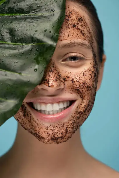 beautiful happy girl with coffee scrub on face, isolated on blue with leaf