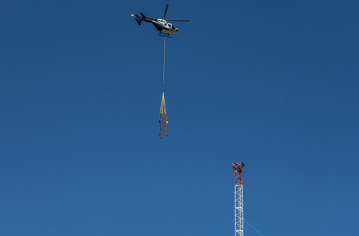Helicopter approaching radio mast with attached component, while workers prepare to assist with positioning