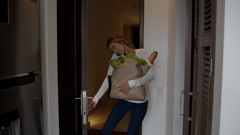 Cheerful young woman arriving home carrying groceries talking on the phone and opening the door