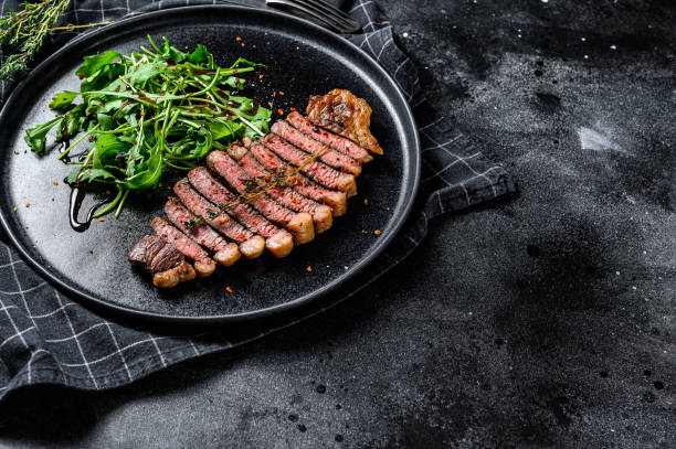 istock Sliced sirloin steak, marbled beef meat with arugula. Black background. Top view. Copy space 1208886962
