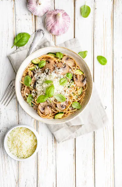 Healthy whole wheat spaghetti with sauteed mushrooms garlic and zucchini, topped with grated cheese and basil leaves on rustic wooden background