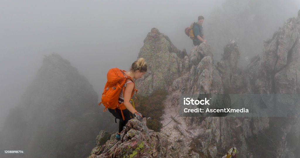 Young woman and young man traverse a ridge on a via ferrata in the fog They are scrambling along a high traverse Cliff Stock Photo