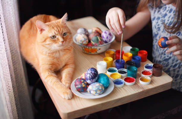 Teen girl paints Easter eggs, her ginger cat lies on the table. - fotografia de stock