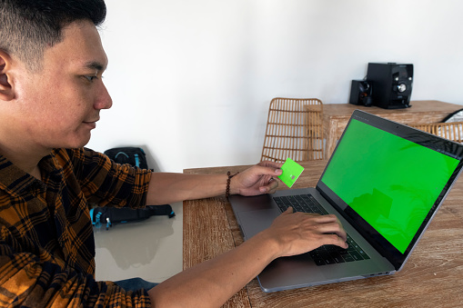 Indonesian young adult man holds a green credit card on his left hand and types on a laptop keyboard with his right one. He is sitting by a wooden table, and looking at the laptop green screen.