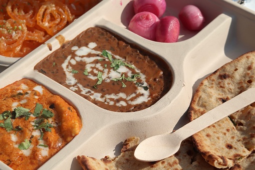 Stock photo showing a disposable plate with compartments filled with traditional chicken tikka masala sauce / paneer butter masala, dal makhani with cream / yoghurt curd topping  as well as herbs and spices, pickled onion accompanied with sweet Indian dessert jalebi, Indian takeaway, being photographed in New Delhi, India. Like school dinners / prison food