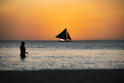 (Selective focus) Stunning view of a boat sailing during a beautiful sunset in the background and the silhouette of a blurred person swimming in the foreground. White Beach, Boracay Island, Philippines.