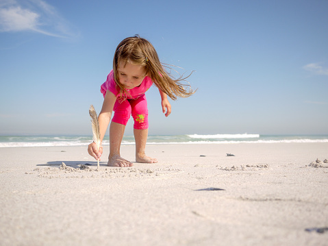 Horizontal View of Beautiful Little Girl Drawing with Feather in the Sand on Beach against Seascape with Clear Sky