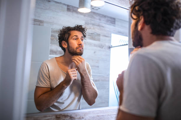 istock Reflection of handsome man with beard looking at mirror and touching face in bathroom grooming. Man putting skincare facial treatment cream on face. 1208975297