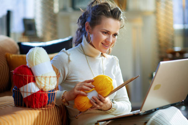 smiling elegant woman with yarn learn how to knit smiling elegant woman in white sweater and skirt with basket with knitting yarn and needles learn how to knit using website on a laptop at modern home in sunny winter day. knitting photos stock pictures, royalty-free photos & images