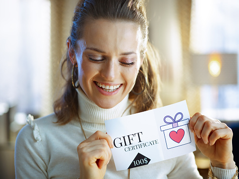 smiling middle age woman in white sweater and skirt with gift certificate at modern home in sunny winter day.