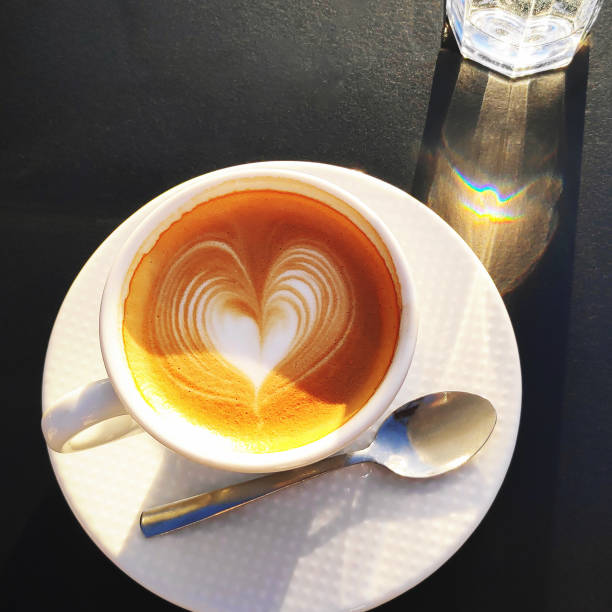 Hot aroma cappuccino or latte coffee in white cup with art on black table in sunlight. Top view.