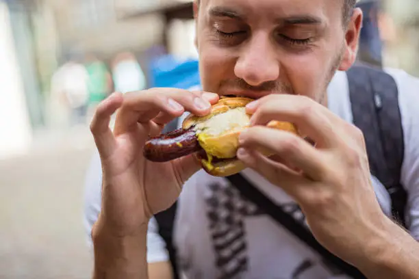 Hungry young male tourist eating German street food, bratwurst at the city street