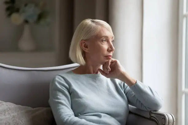 Pensive elderly woman sit on couch look in window distance thinking remembering past good days, thoughtful middle-aged senior female feel distressed lost in thoughts pondering or planning