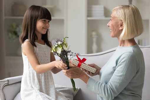 Loving cute small preschooler girl make surprise give gift box and beautiful bouquet to excited senior grandmother, caring little child granddaughter congratulate happy grandparent with anniversary