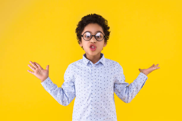 Curly smart clever african american boy in blue shirt and round glasses on yellow background. Black dark skinned cute child is talking, discussing and gesturing. Emotional portrait concept.