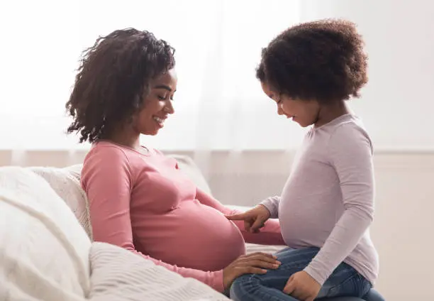 Photo of Curious little black girl touching her pregnant mother's belly at home