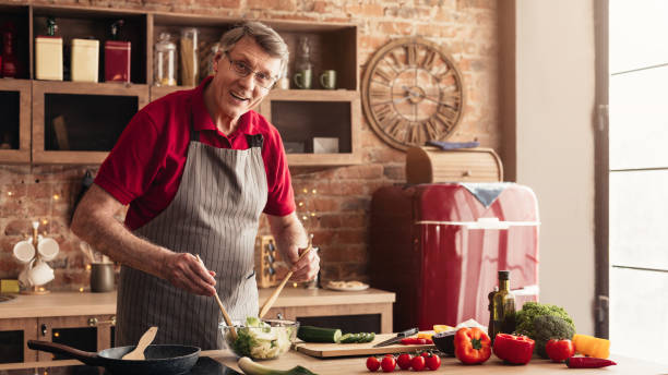 istock Cheerful senior man in apron cooking fresh vegetable salad in kitchen 1208995325