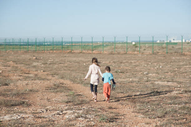 2人の小さな子供の貧しい難民家族の女の子と男の子の不法移民問題は、かみそり有刺鉄線で高いフェンスで州境に向かって熱い砂漠を裸足で歩いています - 孤児 ストックフォトと画像