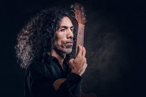 Portrait of attractive curly man with guitar at dark photo studio.