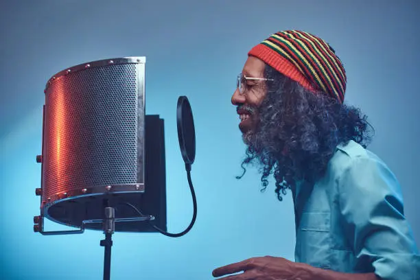 African Rastafarian singer male wearing a blue shirt and beanie emotionally writing song in the recording studio. Isolated on a blue background.