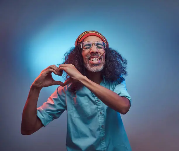 African Rastafarian smokes weed and showing heart symbol and shape with hands. Isolated on a blue background.
