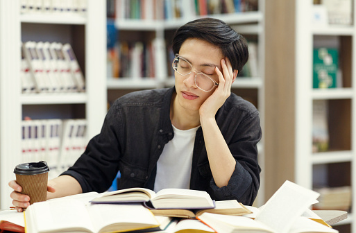 Exhaustion Concept. Sleepy korean guy in glasses napping in library, leaning on elbow, holding cup of coffee