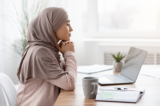Business Planning. Profile portrait of thoughtful arabic female entrepreneur sitting at workplace in office and looking at laptop screen, side view