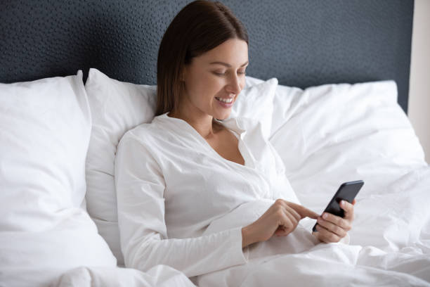istock Smiling woman using phone, sitting in comfortable bed under blanket 1209008672