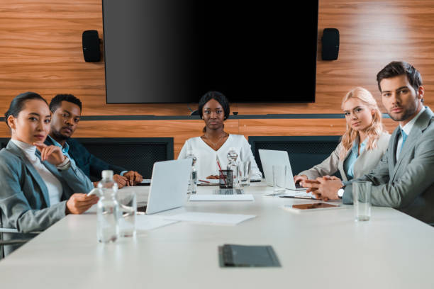 istock young, confident multicultural businesspeople sitting in conference hall with lcd screen on wall 1209011972