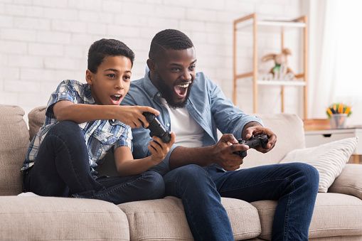 Leisure With Dad. Cheerful Black Father And Son Competing With Each Other In Video Games, Using Joysticks, Having Fun At Home, Free Space